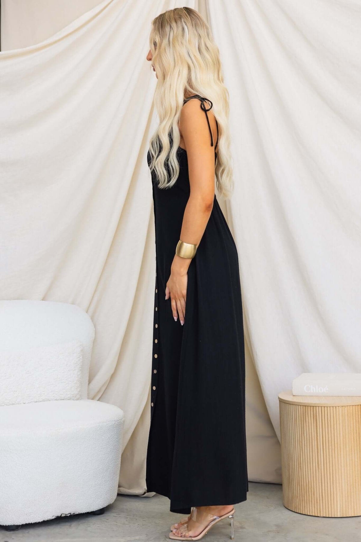 Side view of a woman in a black Damico maxi dress with tie straps and faux buttons.