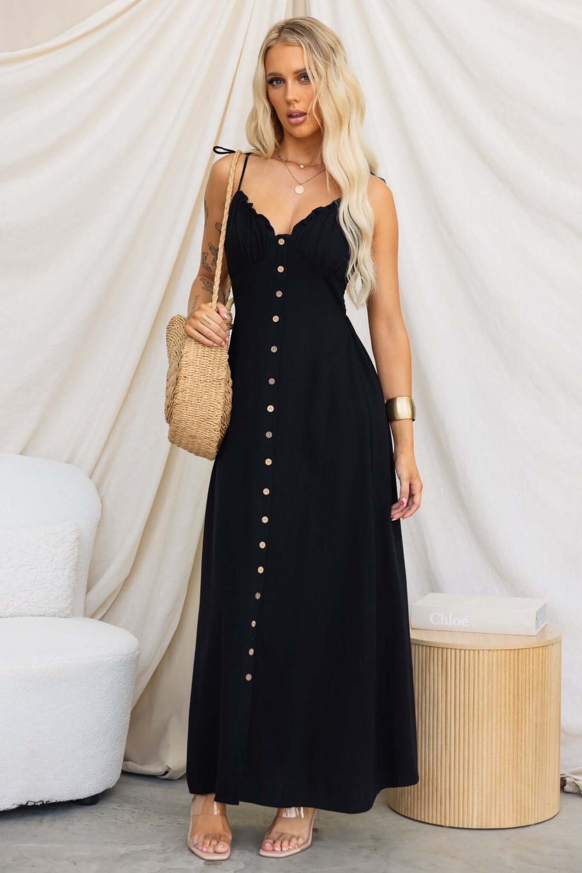 Model wearing Damico Maxi Dress with tie straps and faux buttons, styled with a straw bag and sitting on a modern chair.