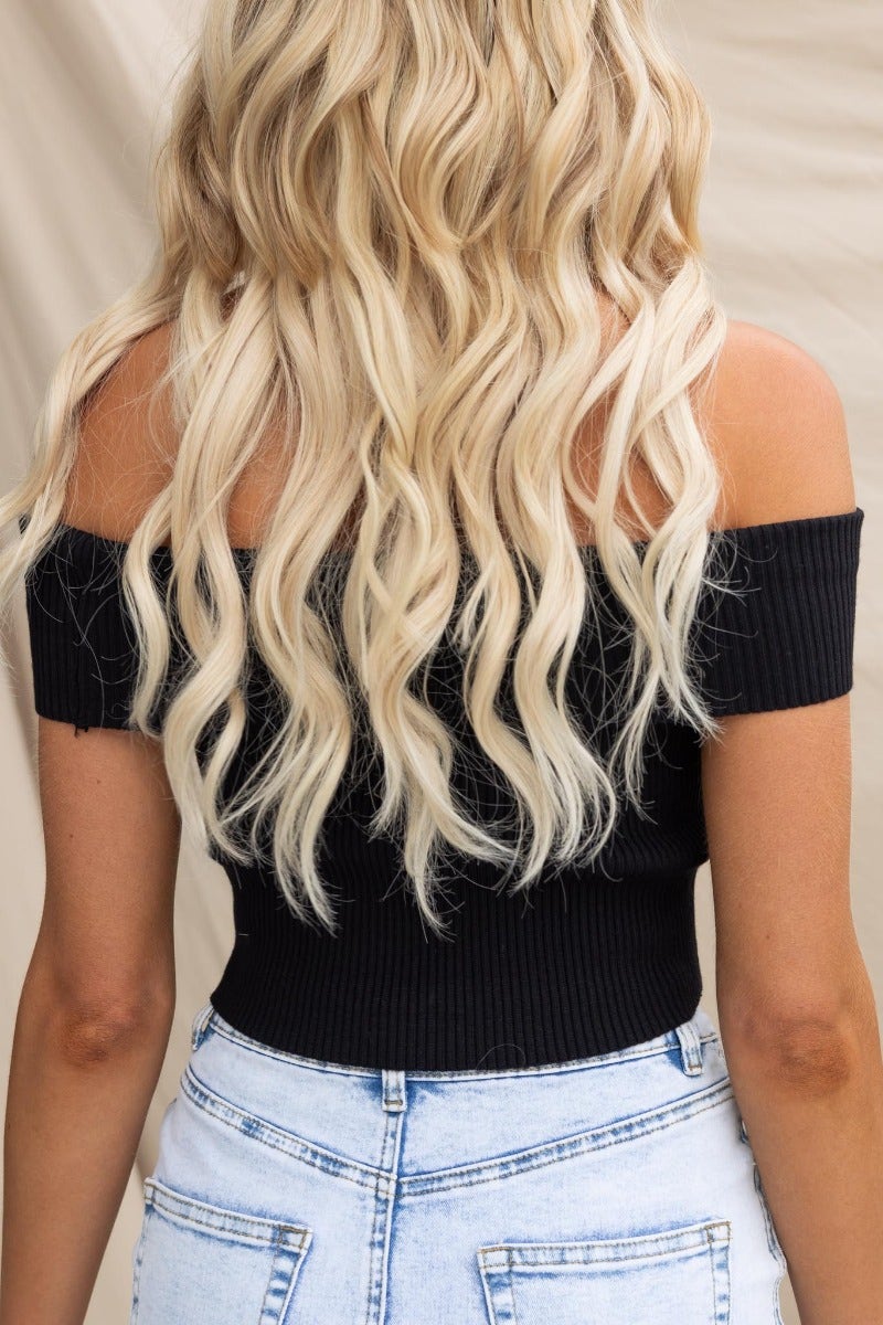Back view of a woman wearing a black ribbed off-shoulder top, styled with wavy blonde hair and high-waisted denim shorts.