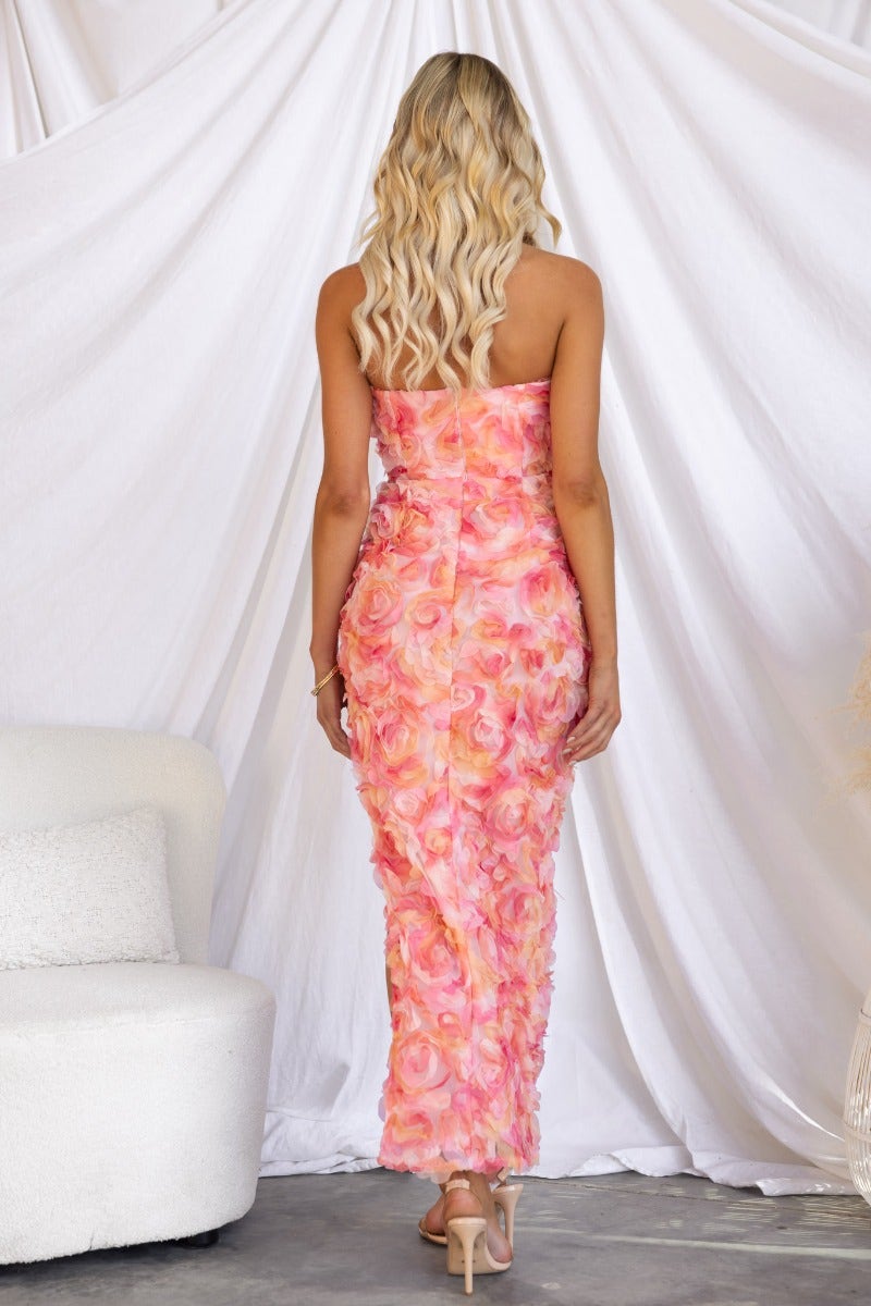 Back view of a model in a strapless lace pink maxi dress with a floral design and asymmetrical hemline.