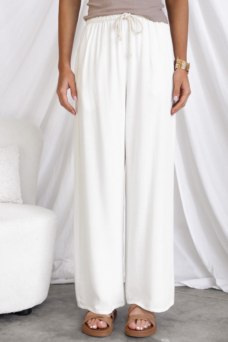 Corella Pants in white with elastic waistband and drawstring, displayed on a model standing on a gray floor.