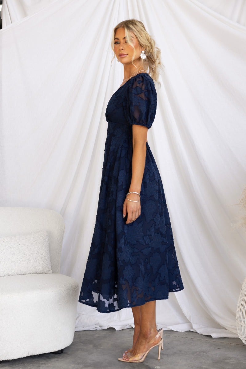 Side view of a woman in a navy embossed midi dress with a V-neckline and puff sleeves, styled with elegant accessories.