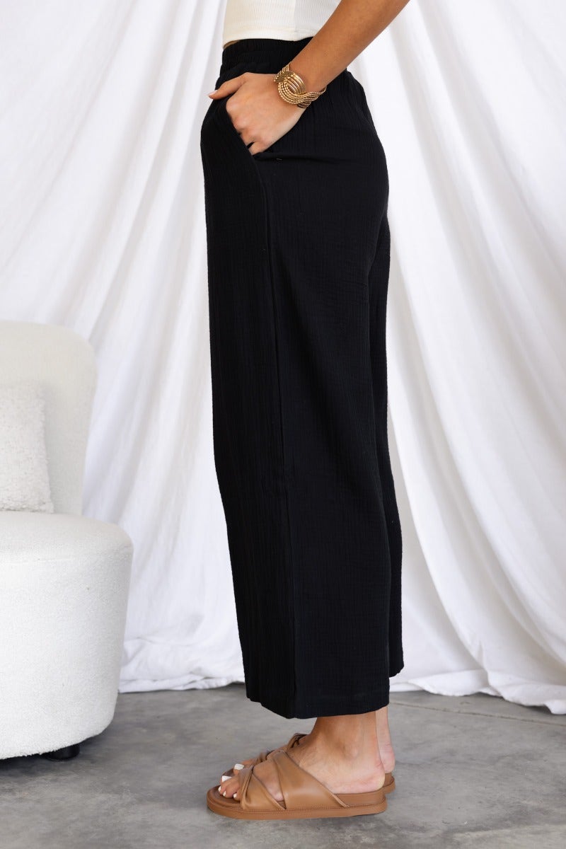 Side view of black cotton blend Eureka pants with elastic waistband and pockets, styled with casual sandals.