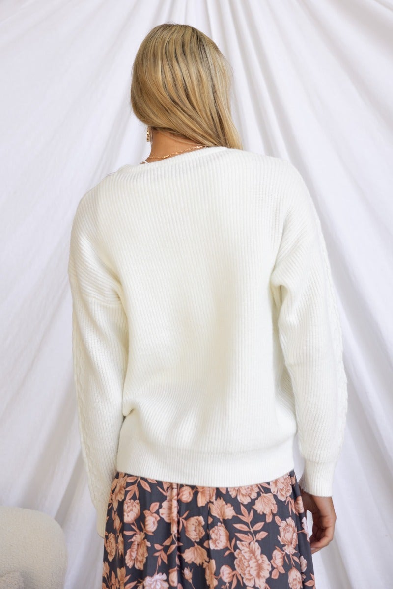 Back view of a woman wearing a white knit Gino Jumper with a floral skirt against a light backdrop.