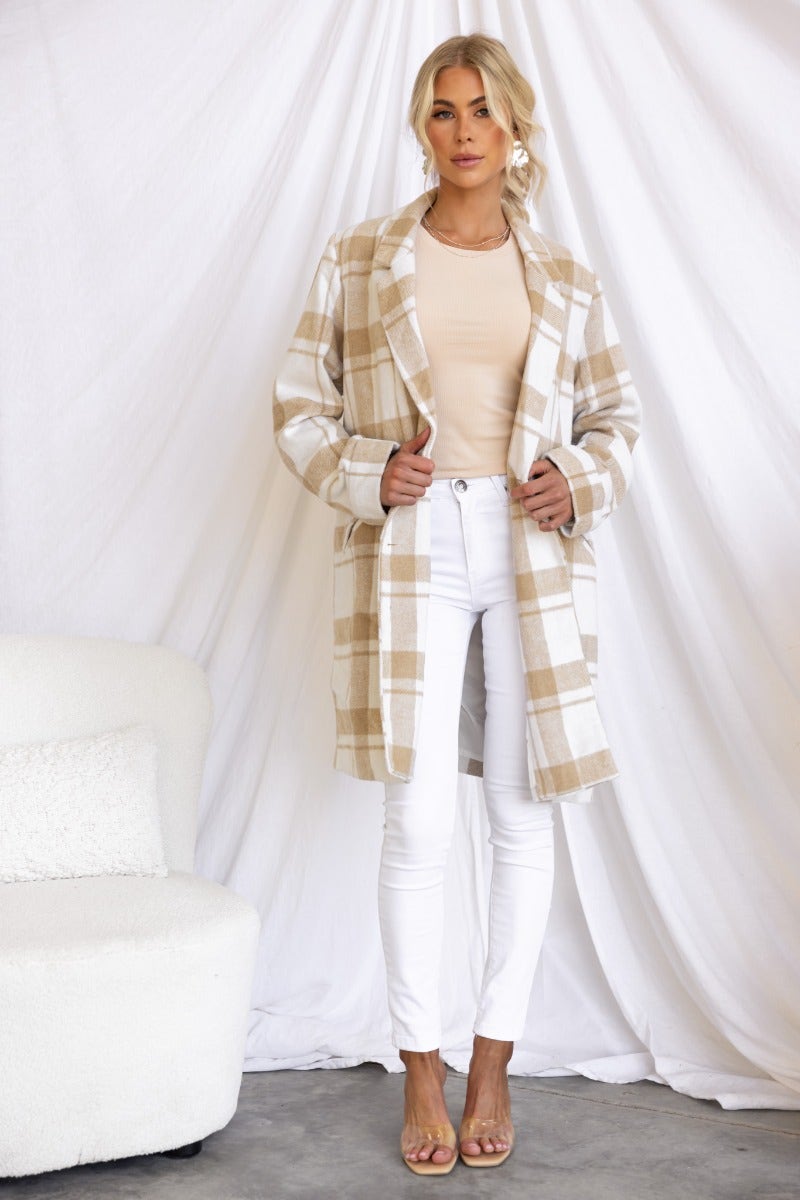 Stylish woman wearing the Sabine plaid coat with beige tones, paired with white jeans and heels, standing against a light backdrop.