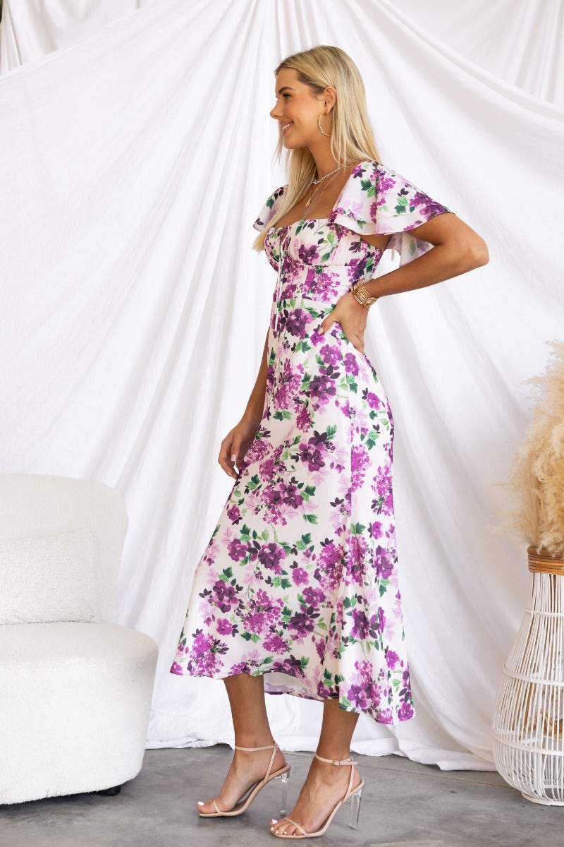 Kamina maxi dress featuring floral print, tie detail at bust, and flutter sleeves, worn by a model in a stylish setting.