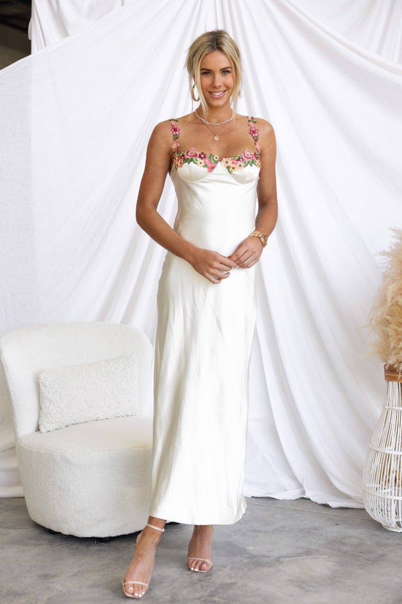 Model wearing a satin Fairy Dust Maxi Dress with embroidered floral trim and adjustable straps, against a soft backdrop.
