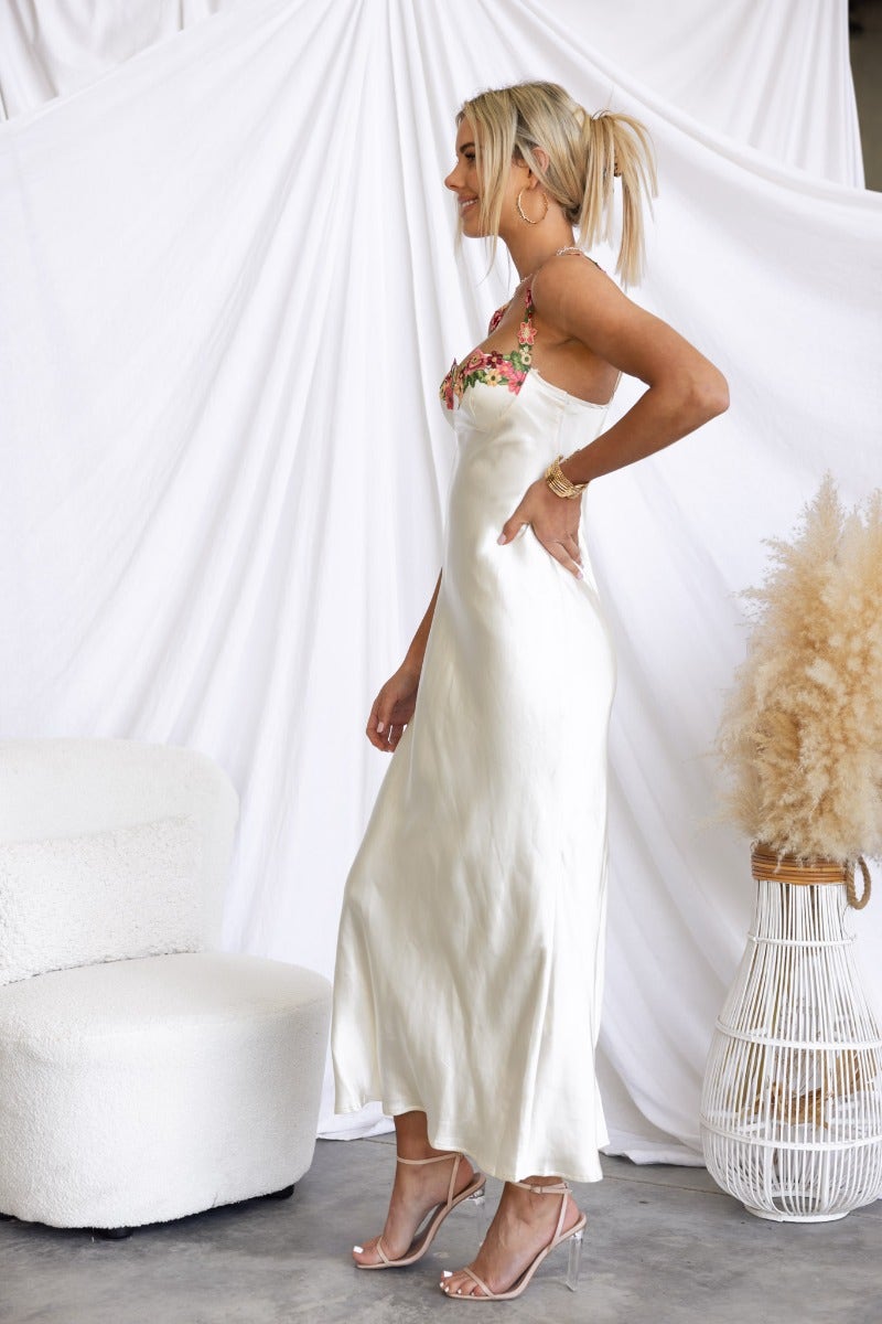 Model wearing Fairy Dust Maxi Dress, a satin floral-embroidered gown with adjustable straps and split, styled elegantly.