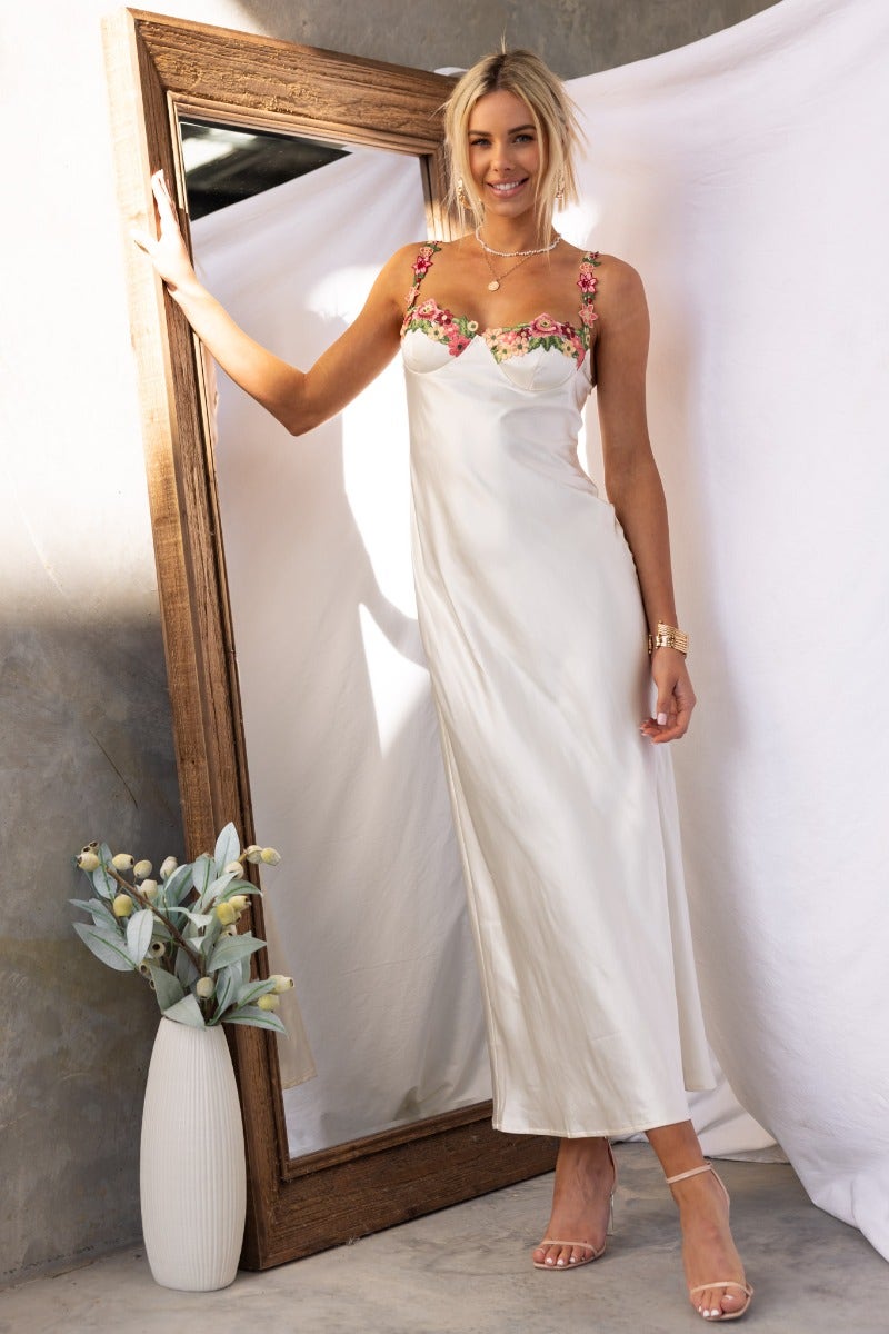Stylish woman in a satin Fairy Dust Maxi Dress with floral trim, posing beside a large mirror in a chic setting.