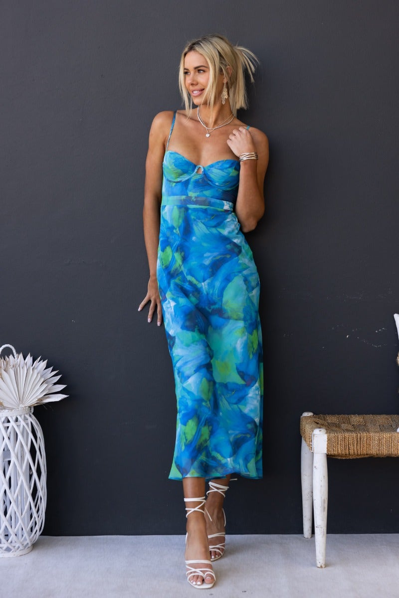 Model wearing a multi-colour print Rhai midi dress with adjustable straps against a dark wall.