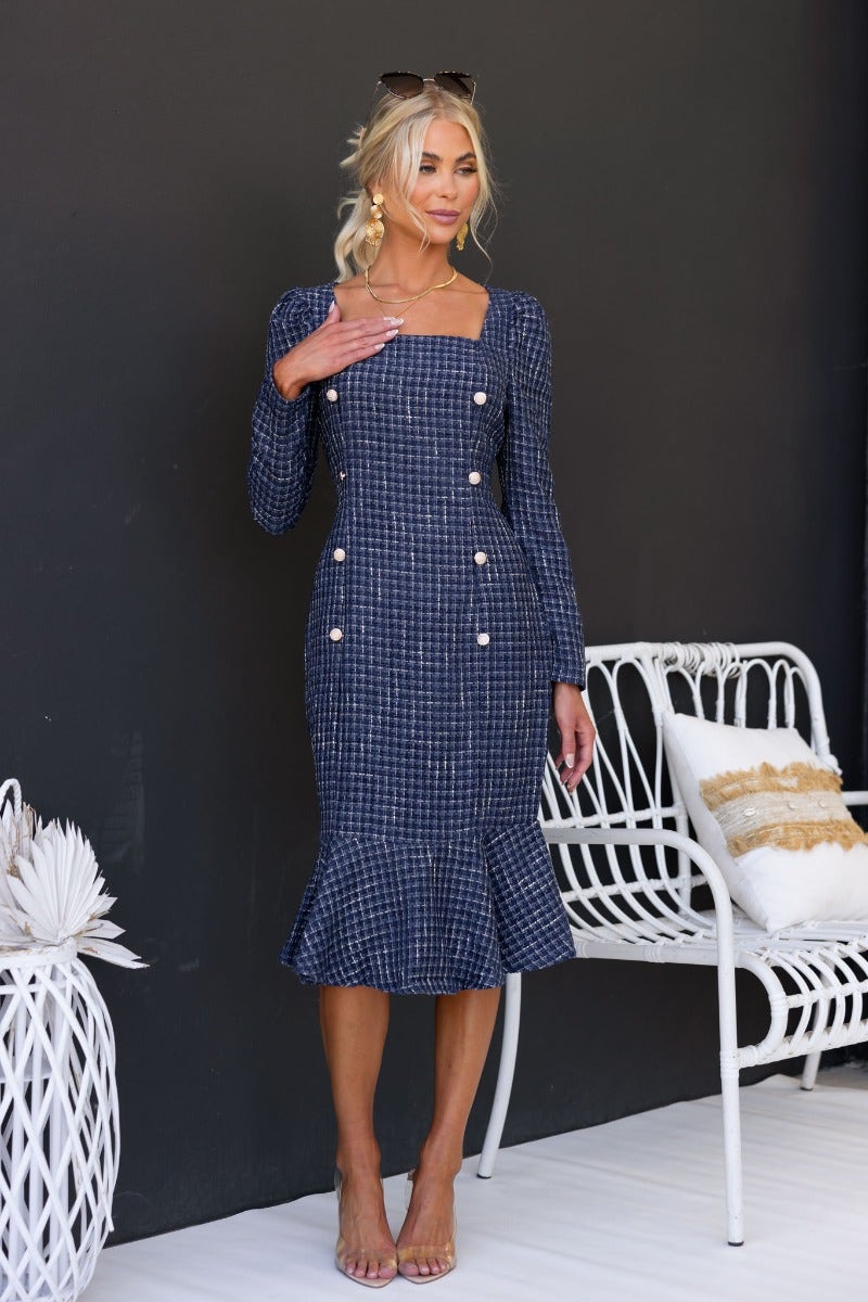 Model wearing a Fine Line Midi Dress with square neckline and gold button detail, standing next to a white chair.