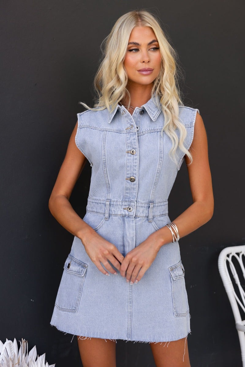 Model wearing Hold Tight Denim Dress, a sleeveless button-up dress with pockets and raw hemline in light denim.