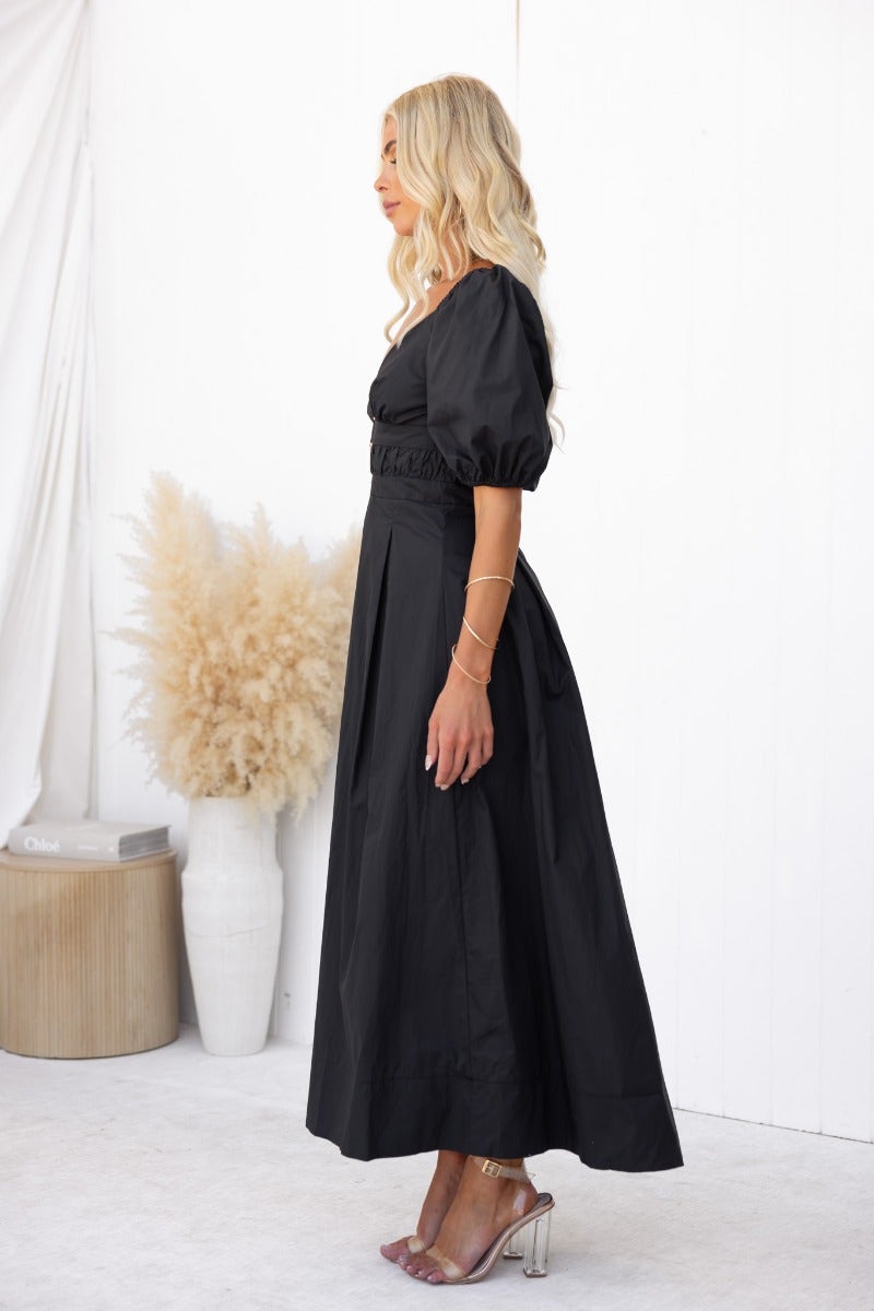 Side view of a model wearing a black cotton maxi dress with puff sleeves and a V-neckline.