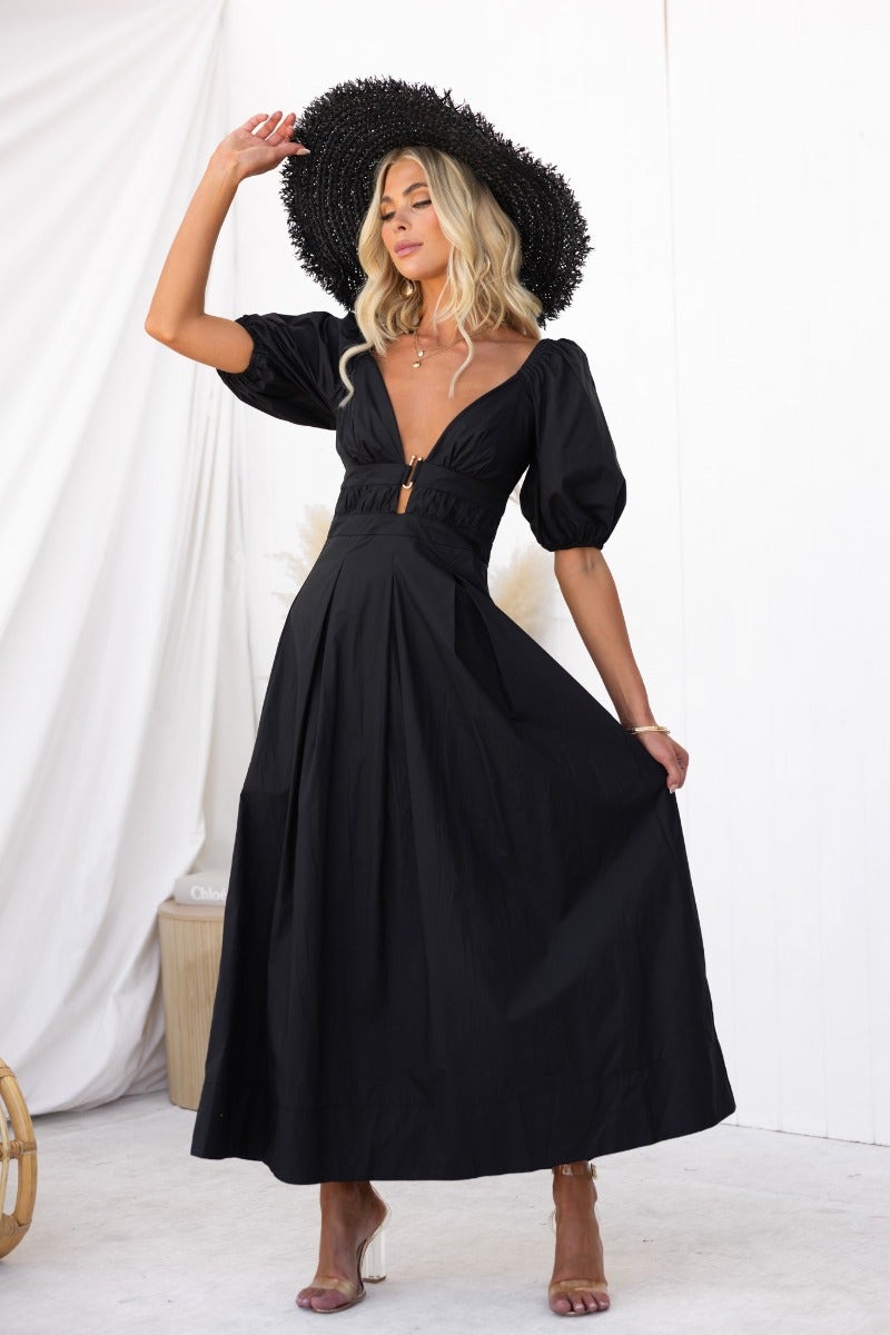 Model wearing The Climb Maxi Dress, a black cotton dress with a V-neckline and gold hardware detail.