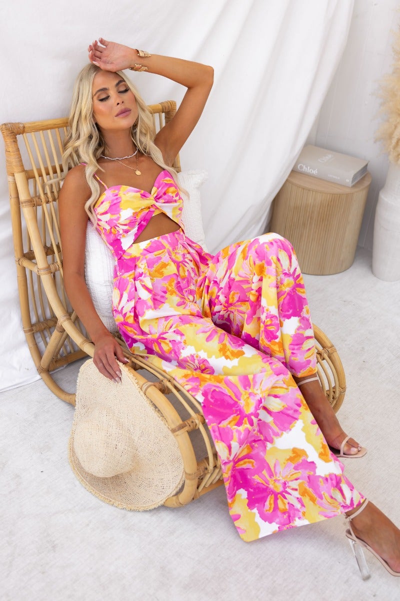 Model in floral Ivy jumpsuit with adjustable straps, twist detail, and pockets, sitting in a rattan chair.