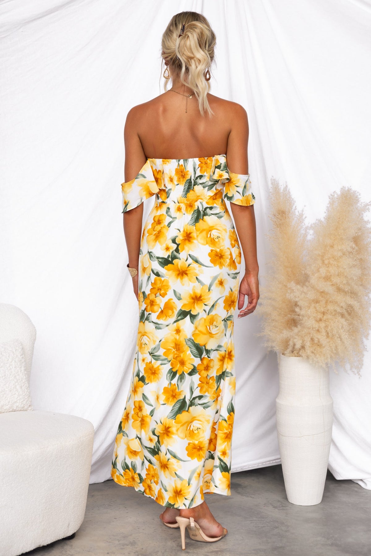 Model wearing a floral off-shoulder maxi dress with yellow flowers from the back, styled with a white backdrop.