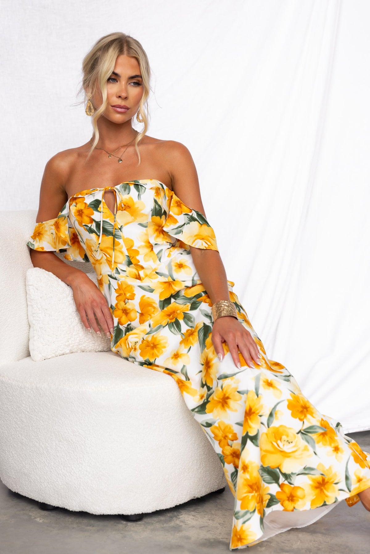 Floral print off-shoulder maxi dress with tie detail and invisible zip, model sitting stylishly on a white chair.