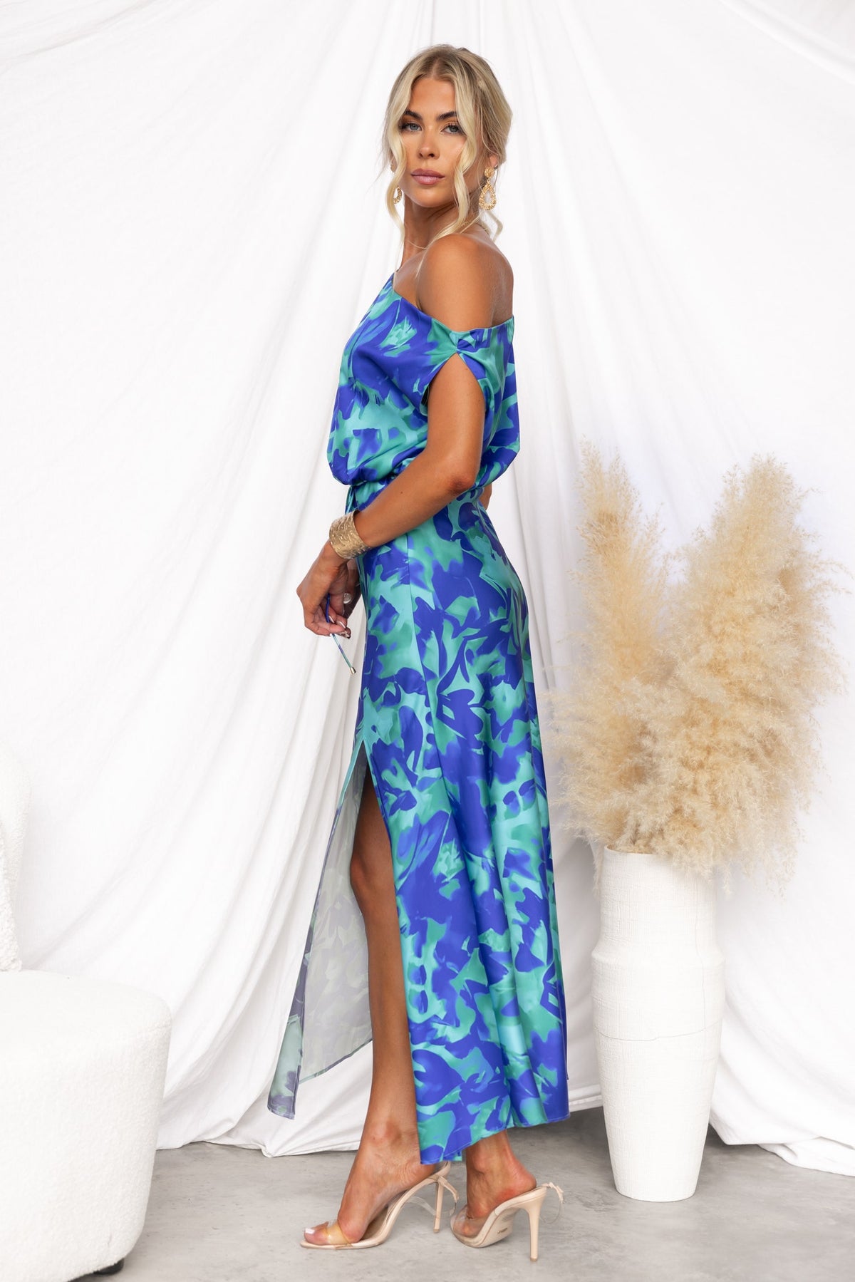 Sicilian Sun maxi dress in blue floral print with asymmetric neckline and side split, worn by a model against a minimalist backdrop.