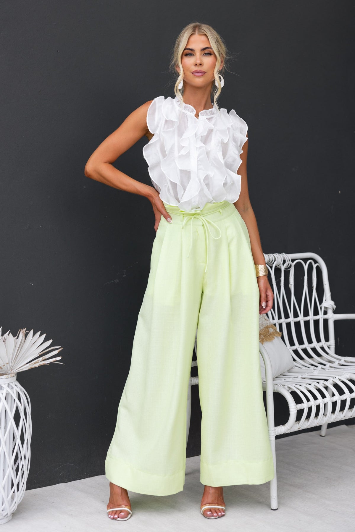 Model wearing a white ruffled button-up top and light yellow wide-leg pants, styled against a modern backdrop.