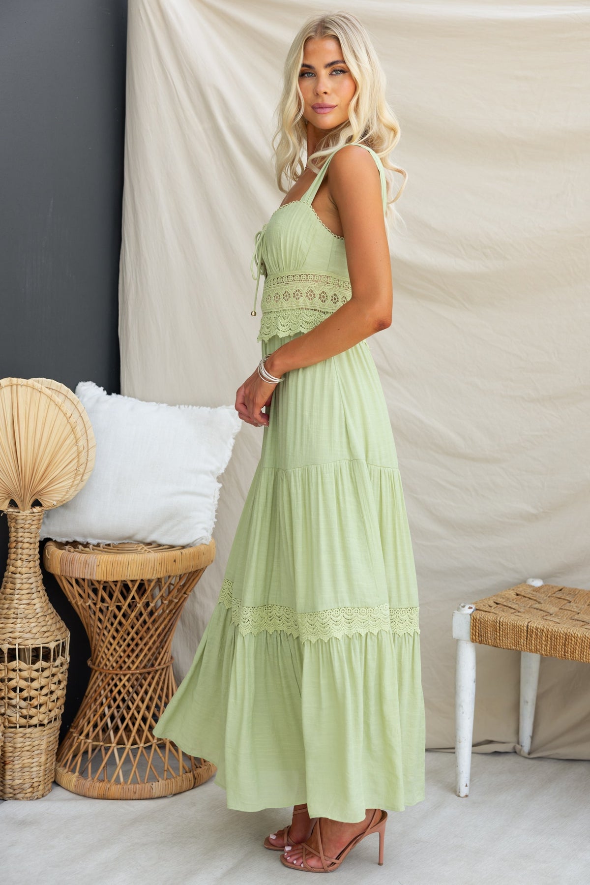 Flaria Maxi Dress in light green featuring tie detail, lace accents at the waist, and flowing skirt, styled with heels.
