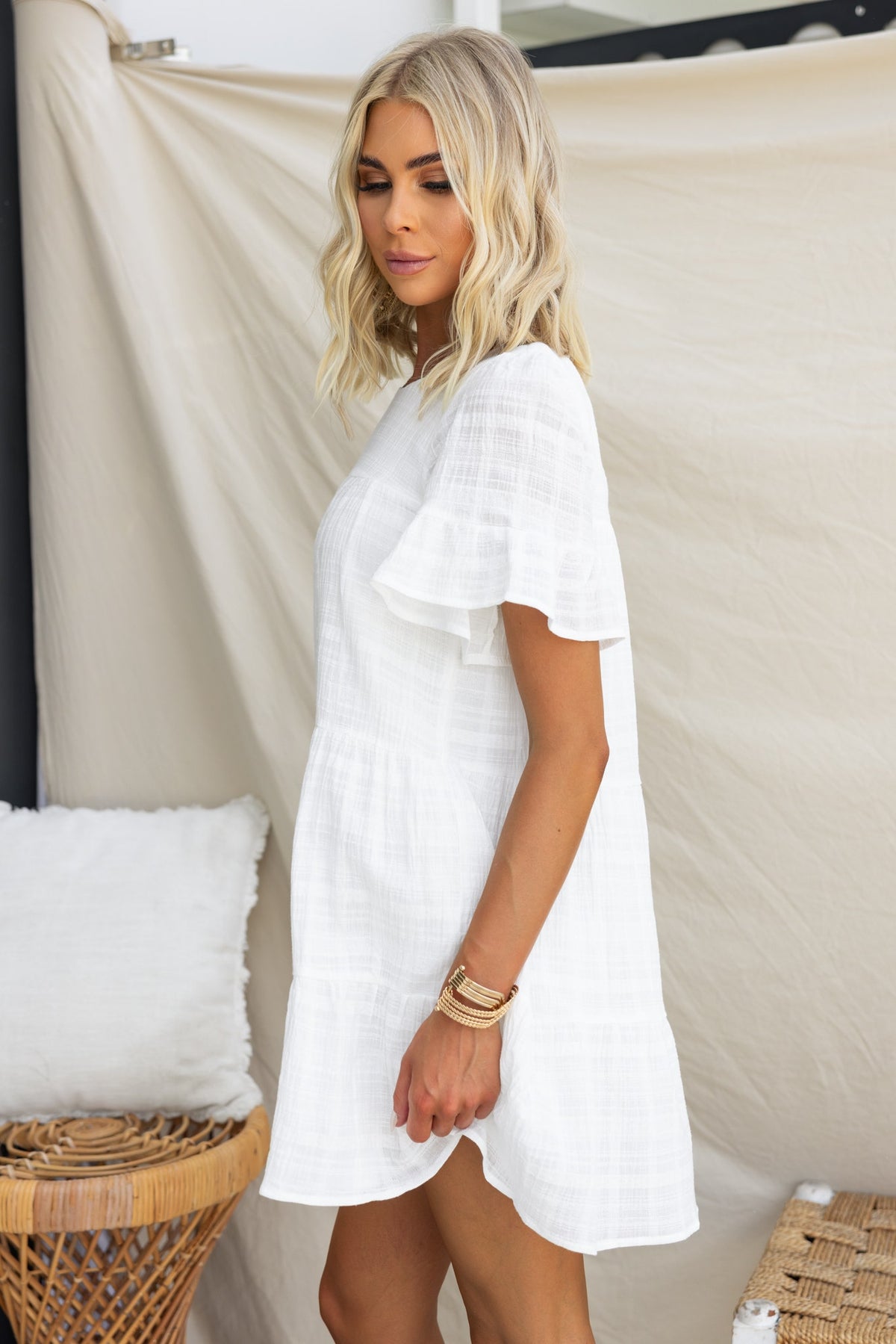 Stylish woman wearing a white Parker Dress with flutter sleeves, standing beside a wicker chair.