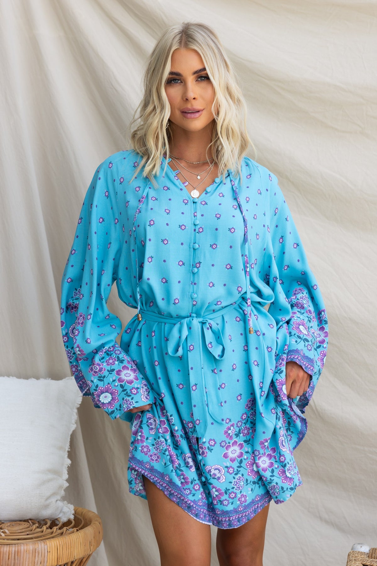 Model wearing Nolla Dress in blue with floral pattern, featuring tie detail, button-up front, and wide sleeves.