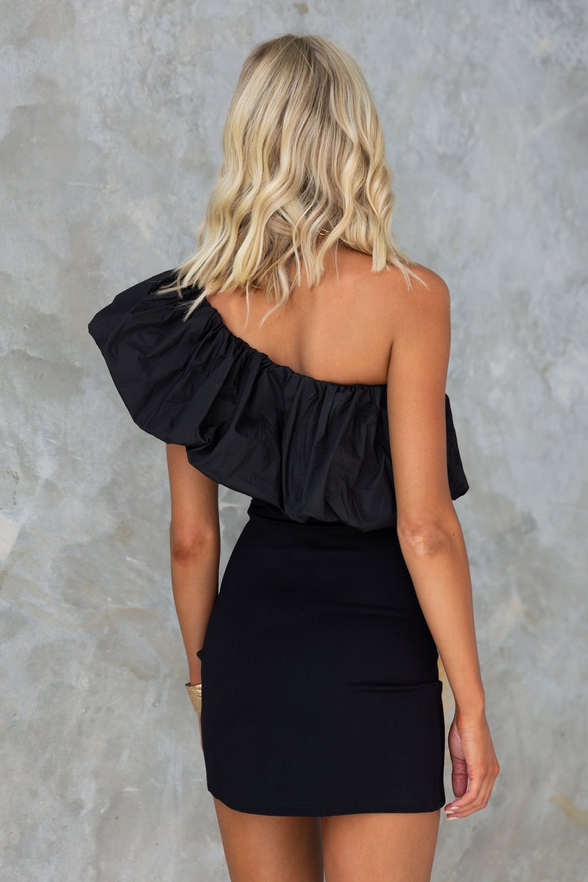 Back view of a model wearing a black one-shoulder Sabre dress with ruffle detail.