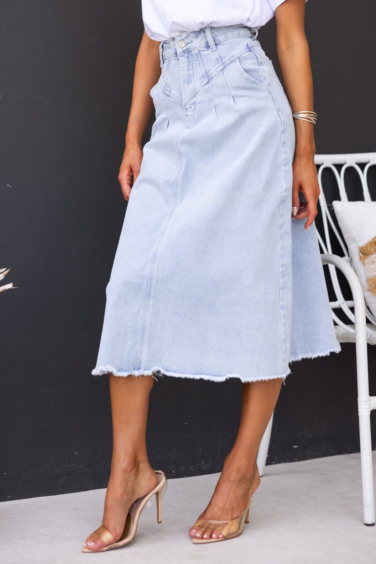 Emera Denim Midi Skirt featuring a mid-high waist, frayed hemline, and front and back pockets, styled with heels.