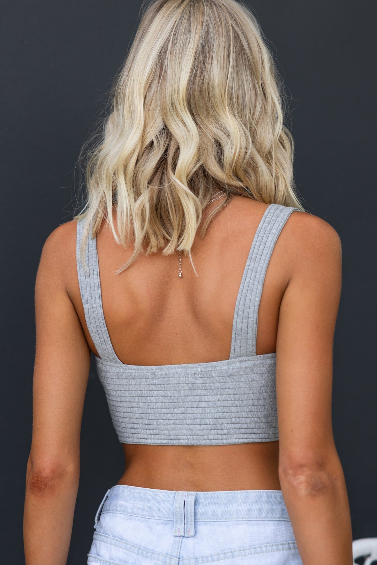Back view of a woman wearing a grey ribbed crop top with wide shoulder straps, styled with light denim shorts.