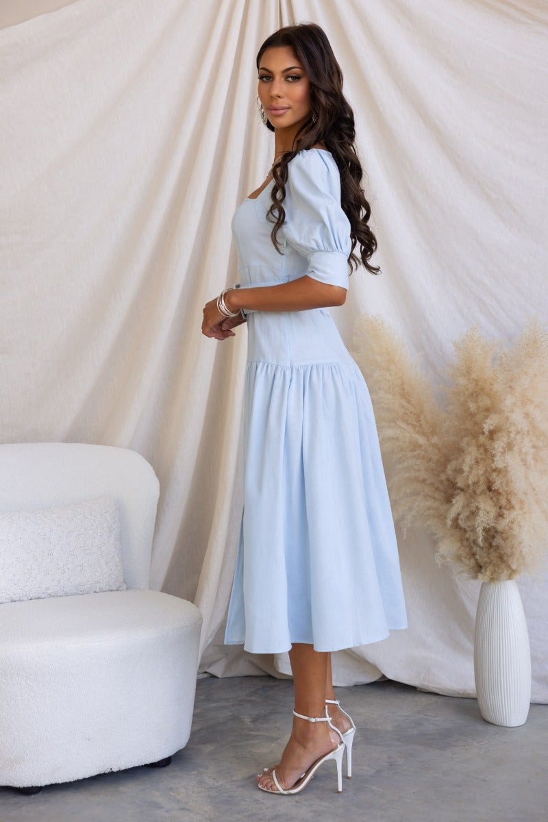Model wearing a light blue linen blend midi dress with square neckline and puff sleeves, showcasing a side split.