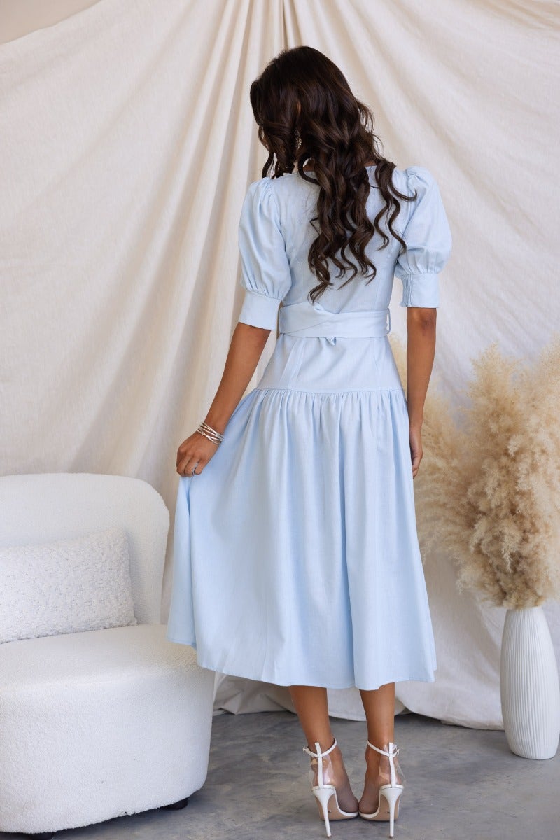 Back view of a woman in a light blue linen blend Innis midi dress with elastic cuffs and removable belt.
