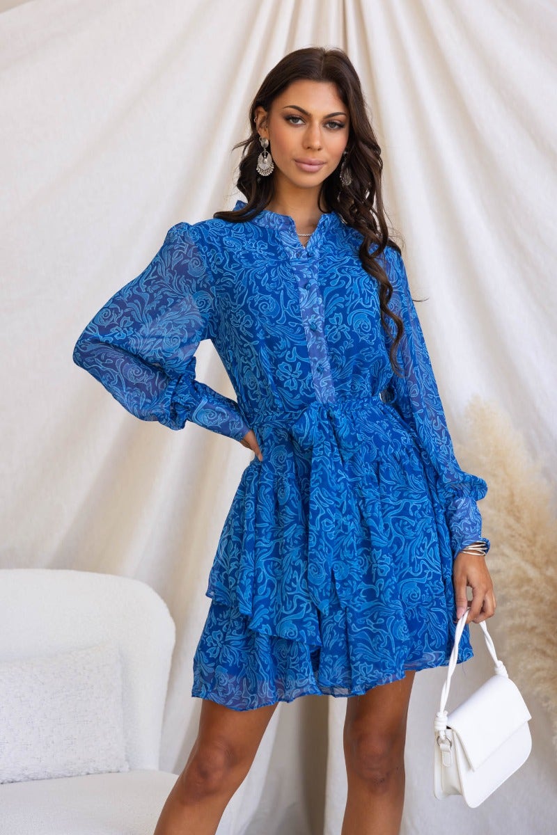 Model wearing a blue Revival Dress with a high neckline, button-up front, and removable belt, styled with a white handbag.