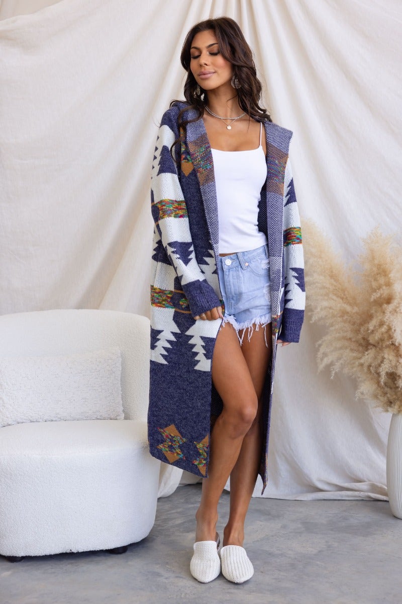 Model wearing a longline navy cardigan with pockets and attached hood, paired with denim shorts and white top.