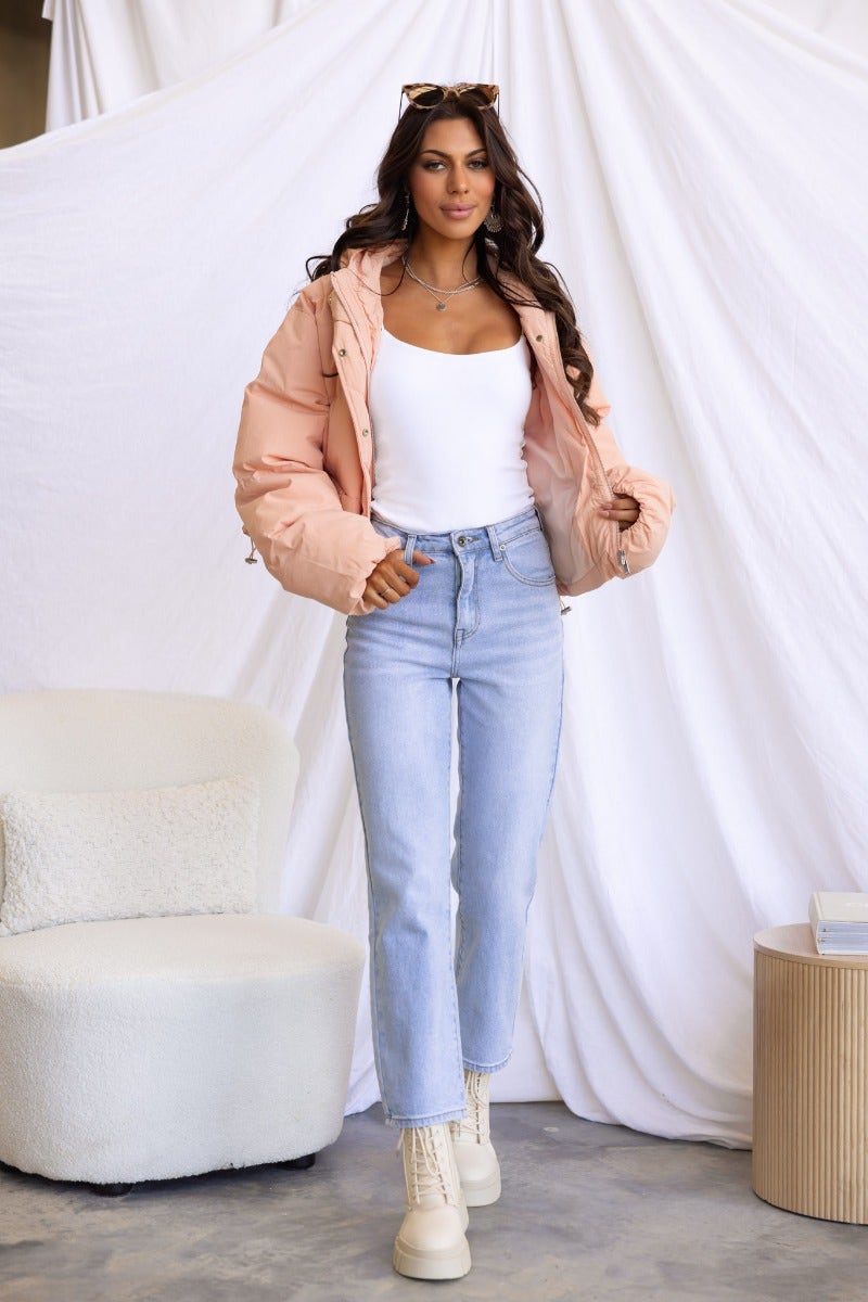 Woman in a pink Vinna Puffer Jacket, white tank top, and light jeans, posing in a stylish indoor setting.