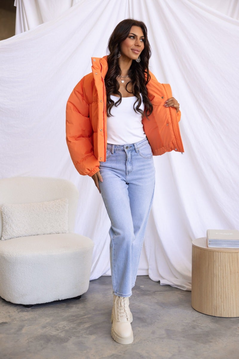 Model wearing an orange Avril puffer jacket, styled with light blue jeans and cream boots, posing in a modern interior.