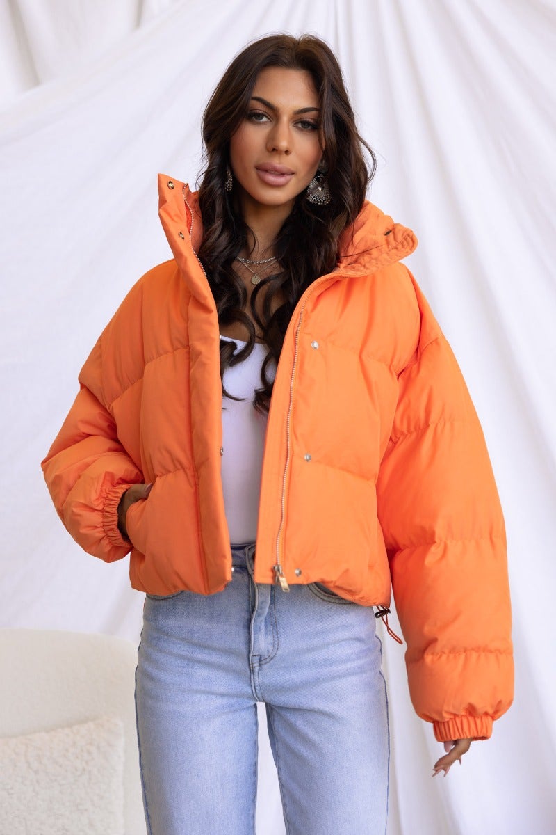 Model wearing an orange Avril Puffer Jacket with high neckline and adjustable drawstring, paired with blue jeans.