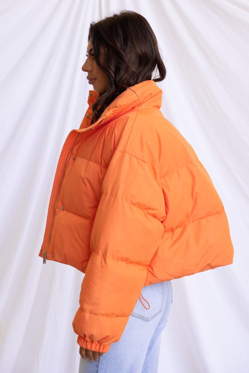 Avril Puffer Jacket in vibrant orange, featuring a high neckline, button and zip closure, and elastic cuffs.