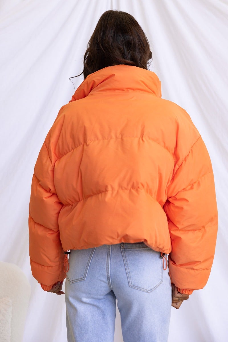 Back view of Avril Puffer Jacket in vibrant orange, featuring high neckline and elastic cuffs.