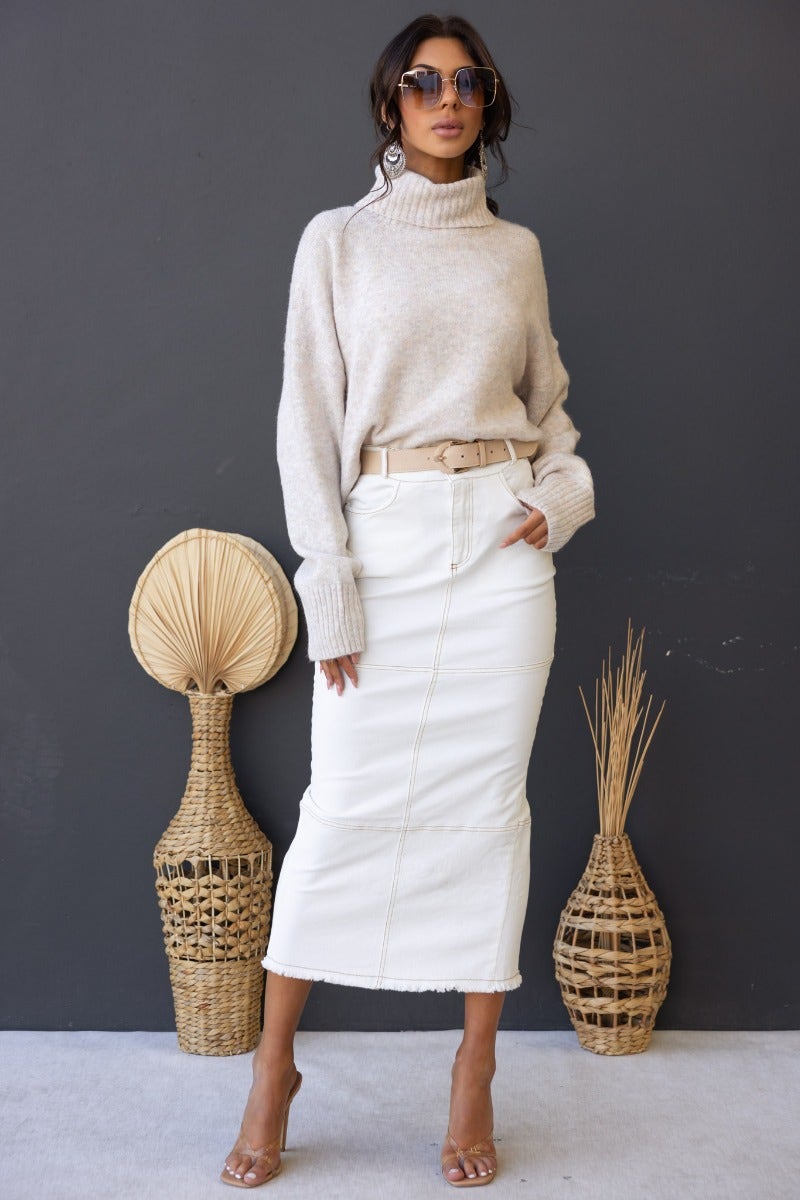 Woman wearing a beige ribbed knit turtle neck jumper paired with a white midi skirt and stylish sunglasses.