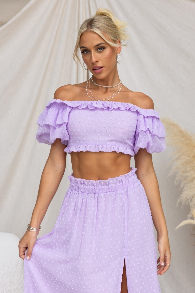 Young woman in a textured lavender off-shoulder top with ruffles, styled with a matching skirt.