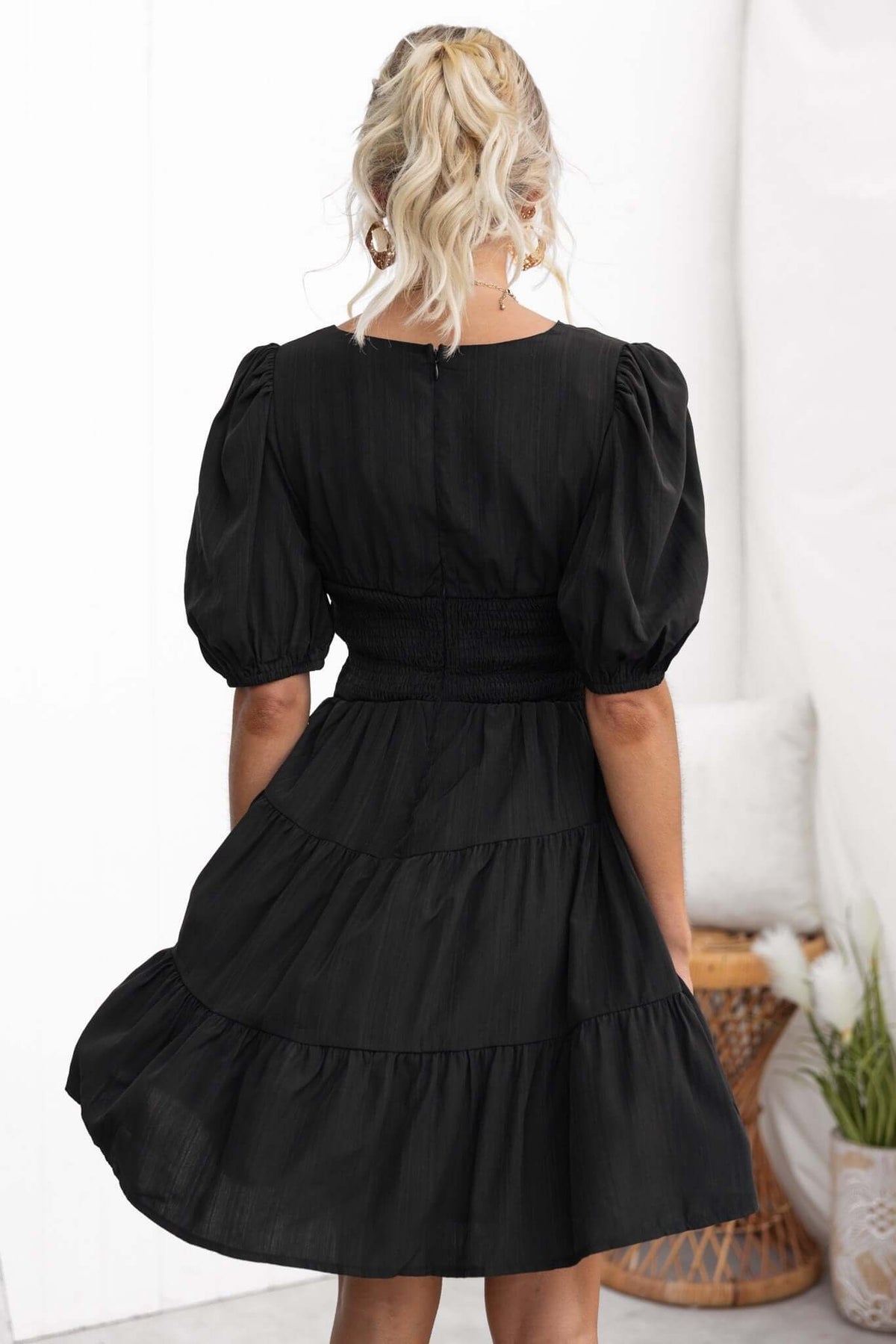 Back view of a black Sakura dress featuring puff sleeves and a tiered skirt, modeled by a woman in a stylish setting.