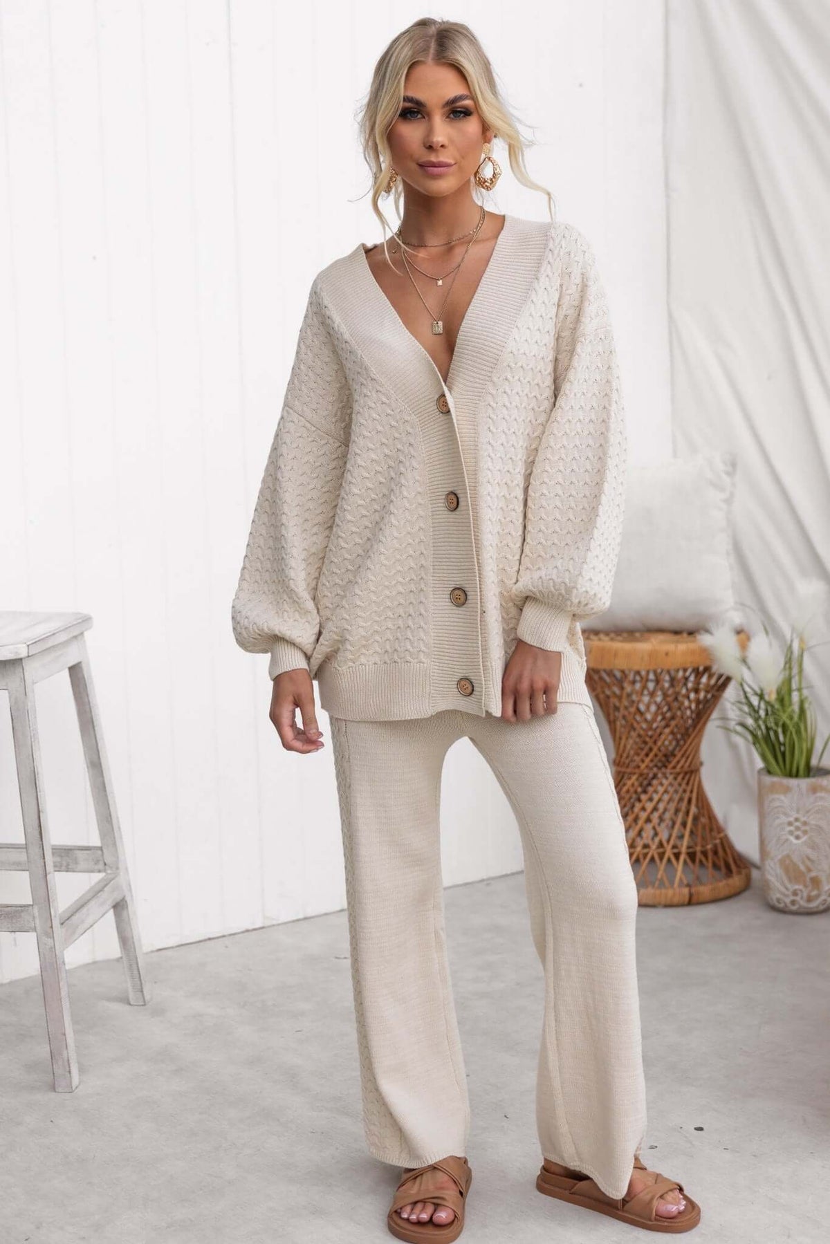 Model wearing a beige knitted cardigan and matching pants, styled with layered necklaces and sandals.