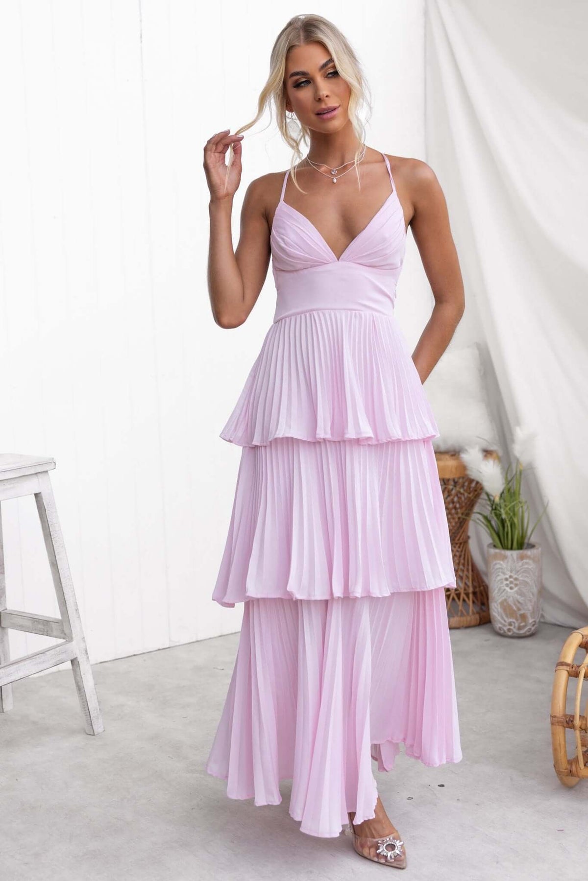 Model wearing a pink Remmy Maxi Dress with pleated ruffle skirt and tie straps in a bright studio setting.