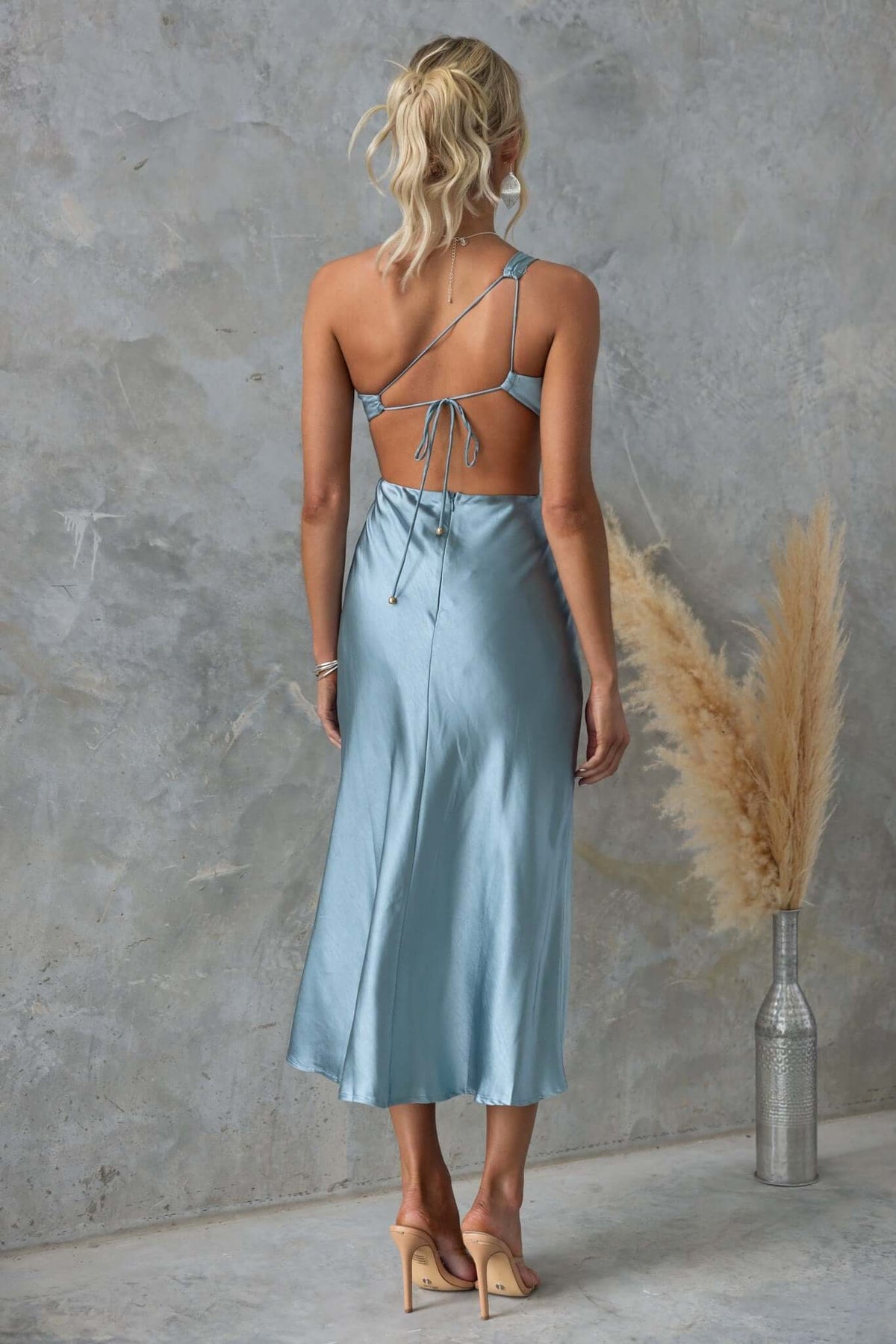 Back view of a silk blue midi dress with tie straps and cut-out waist, styled with nude heels and decorative foliage.
