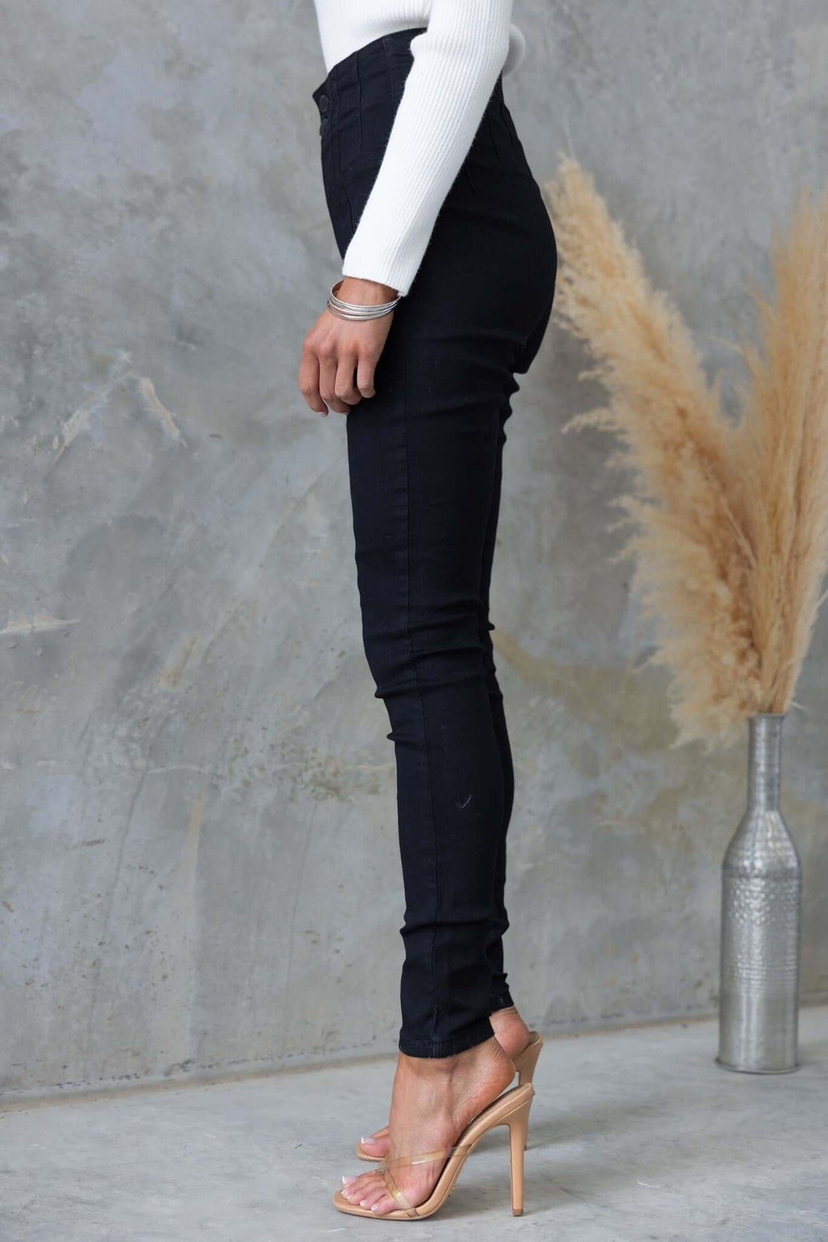 Side view of a model wearing Okara Jeans, black denim, mid-high waist, paired with stylish heels.