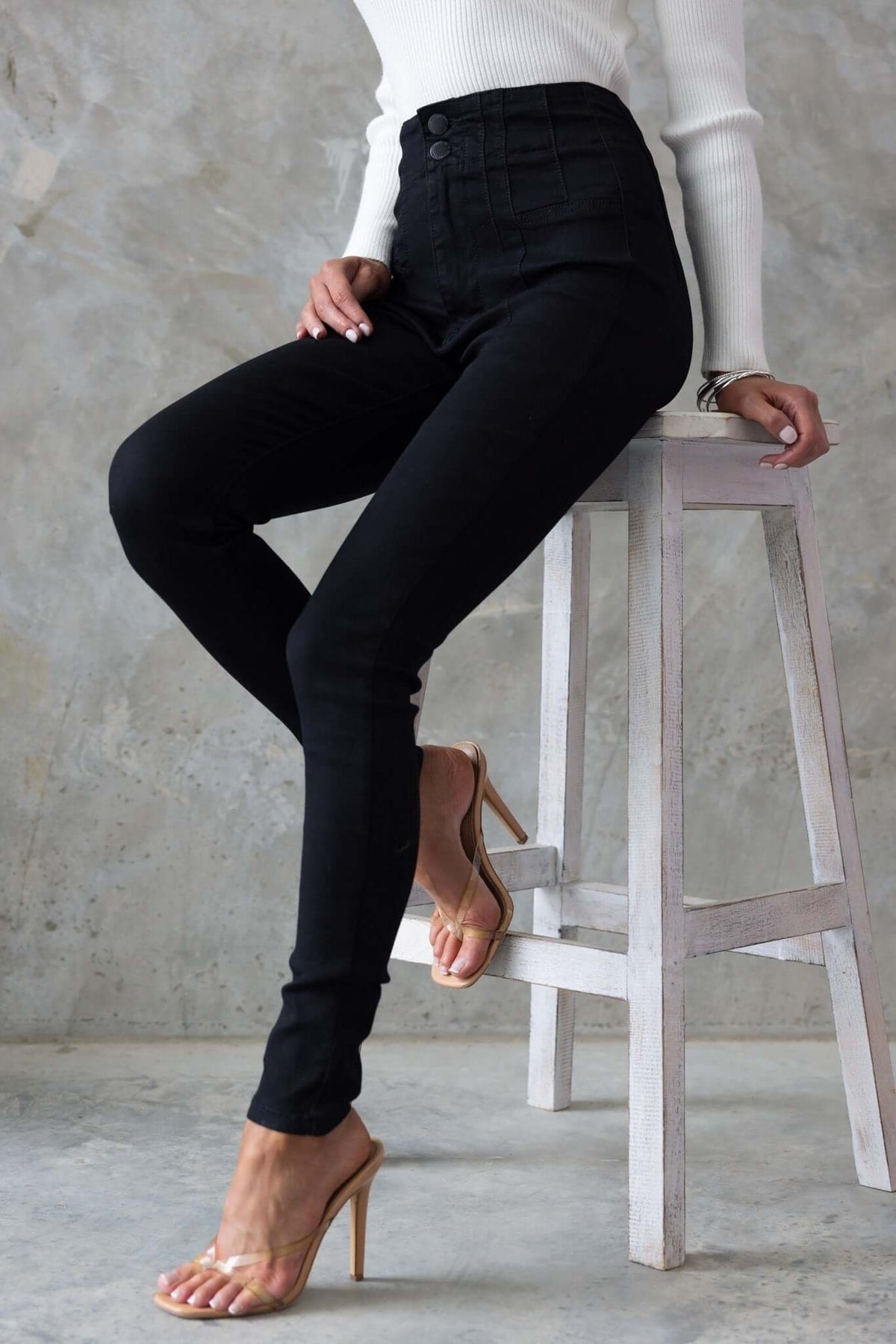 Model wearing black denim Okara jeans with a mid-high waist and a two-button closure, seated on a stool.