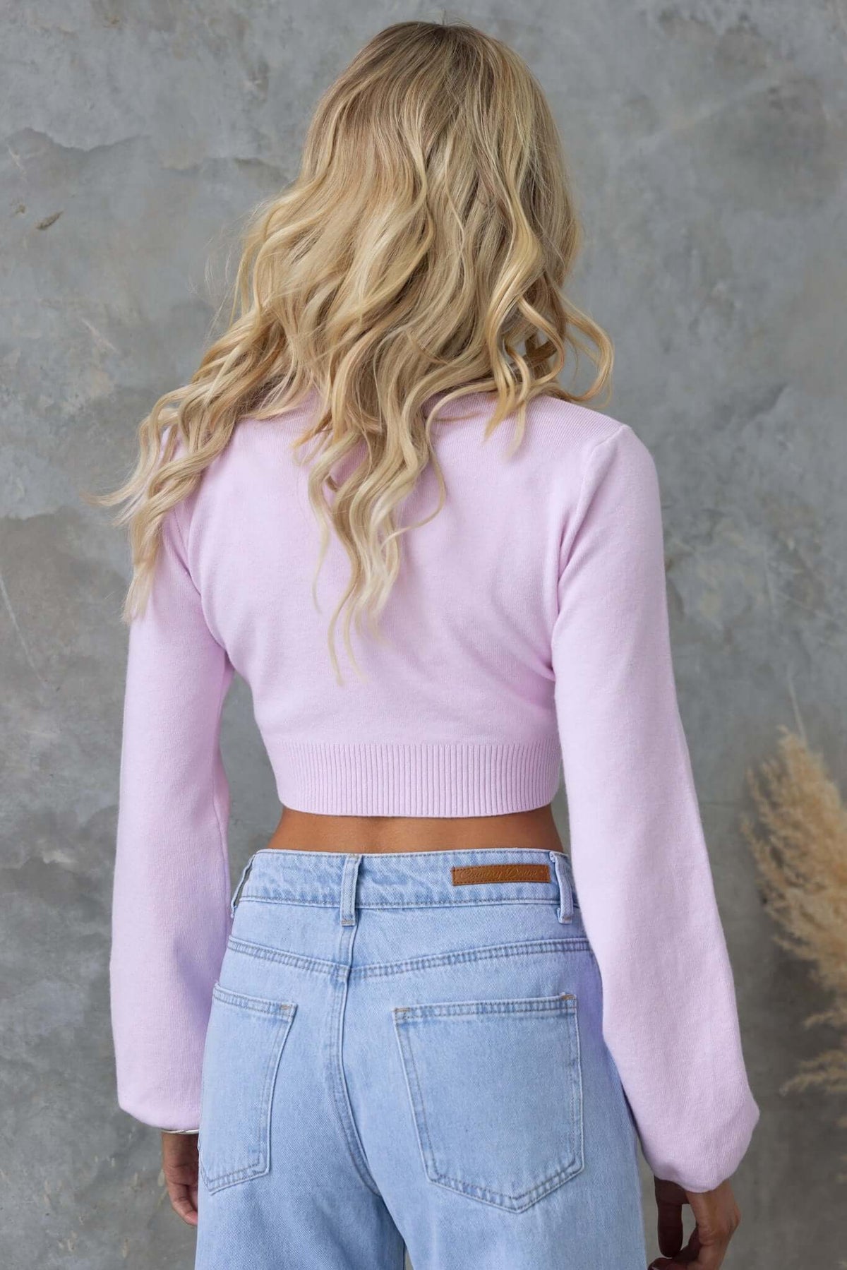Back view of a woman in a pink Sora jumper paired with light blue jeans, showcasing its cropped design and knit material.