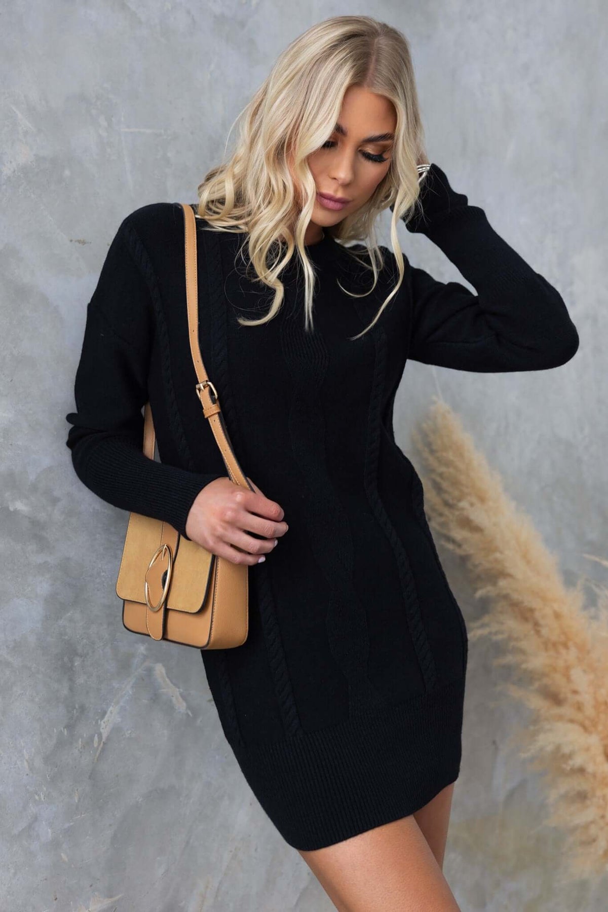 Model wearing Beasley Dress, a black cable knit sweater dress, paired with a tan bag against a textured background.