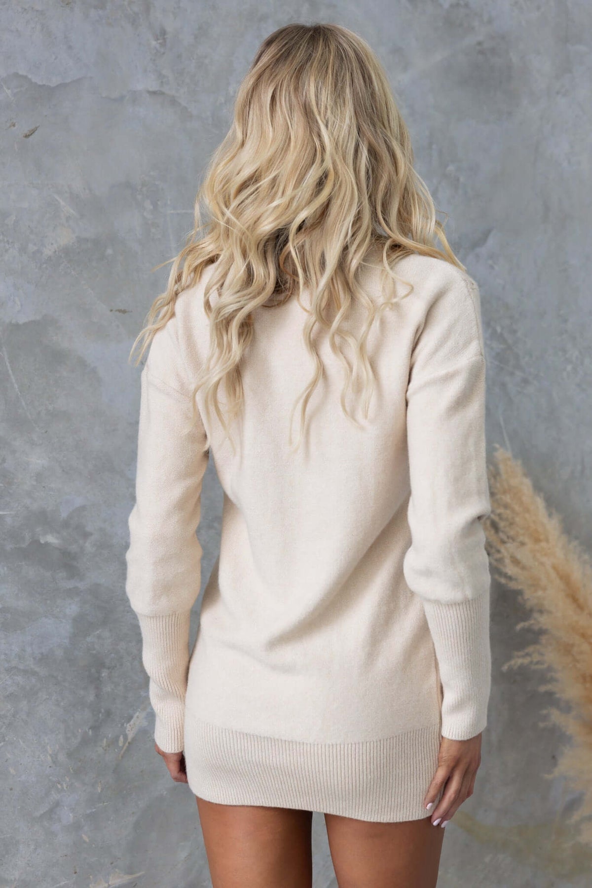 Woman with long blonde hair wearing a beige knit dress, showcasing the back view against a textured background.