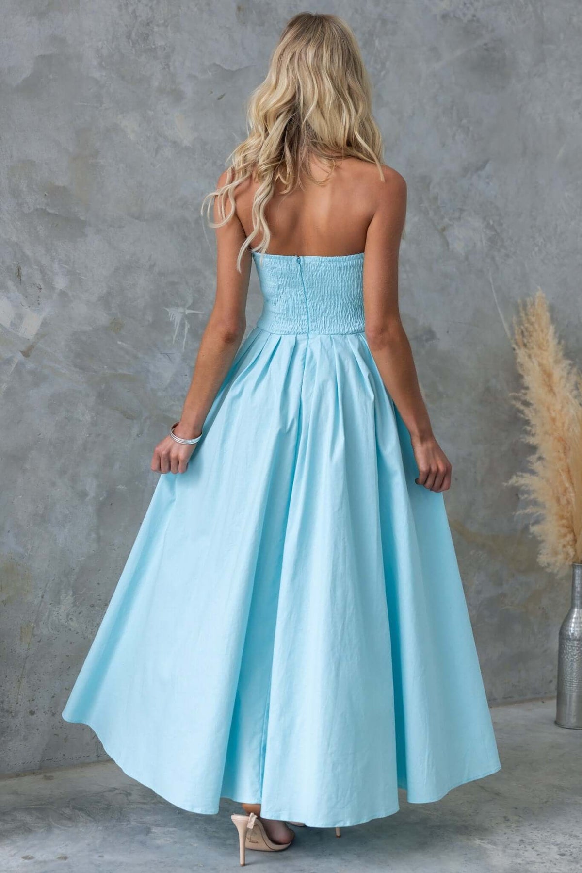 Back view of a light blue strapless maxi dress featuring an asymmetric hem and boning at the bust, styled with elegant hair.